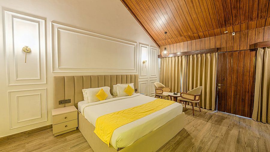 Spacious and elegant cottage room with a large bed covered in white and yellow linens, wooden paneled ceiling, and large windows with thick drapes overlooking tropical greenery.