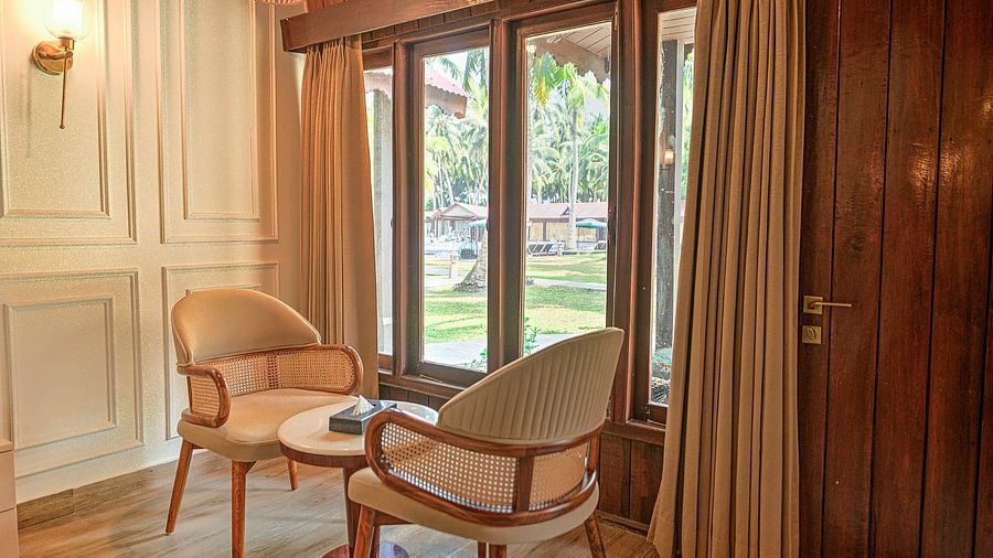 Bright and cozy seating area inside a cottage with two wicker chairs and a small table, large windows offering a view of the tropical surroundings and the beachside.