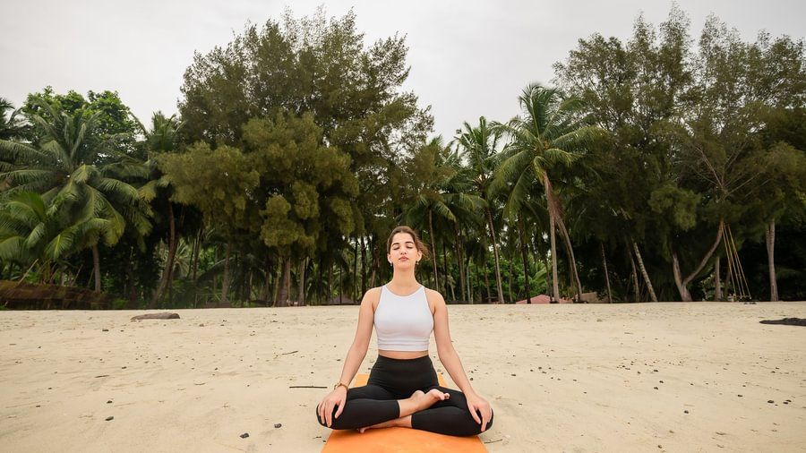 Woman meditating in lotus position on a yoga mat on a beach with a background of dense palm trees.