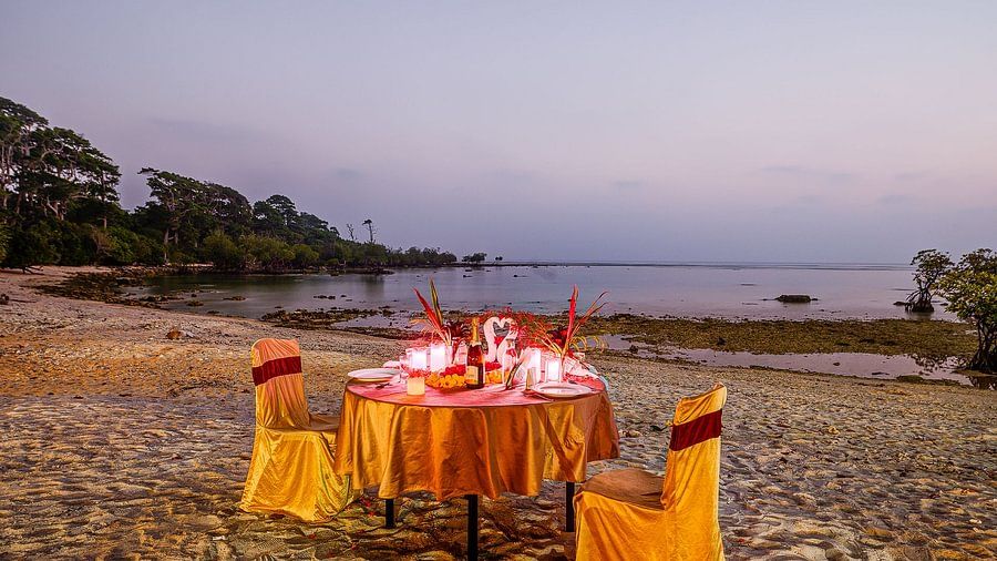 A romantic beach dinner setup with a table covered in a golden cloth, surrounded by candles and with a twilight sky and sea in the background.