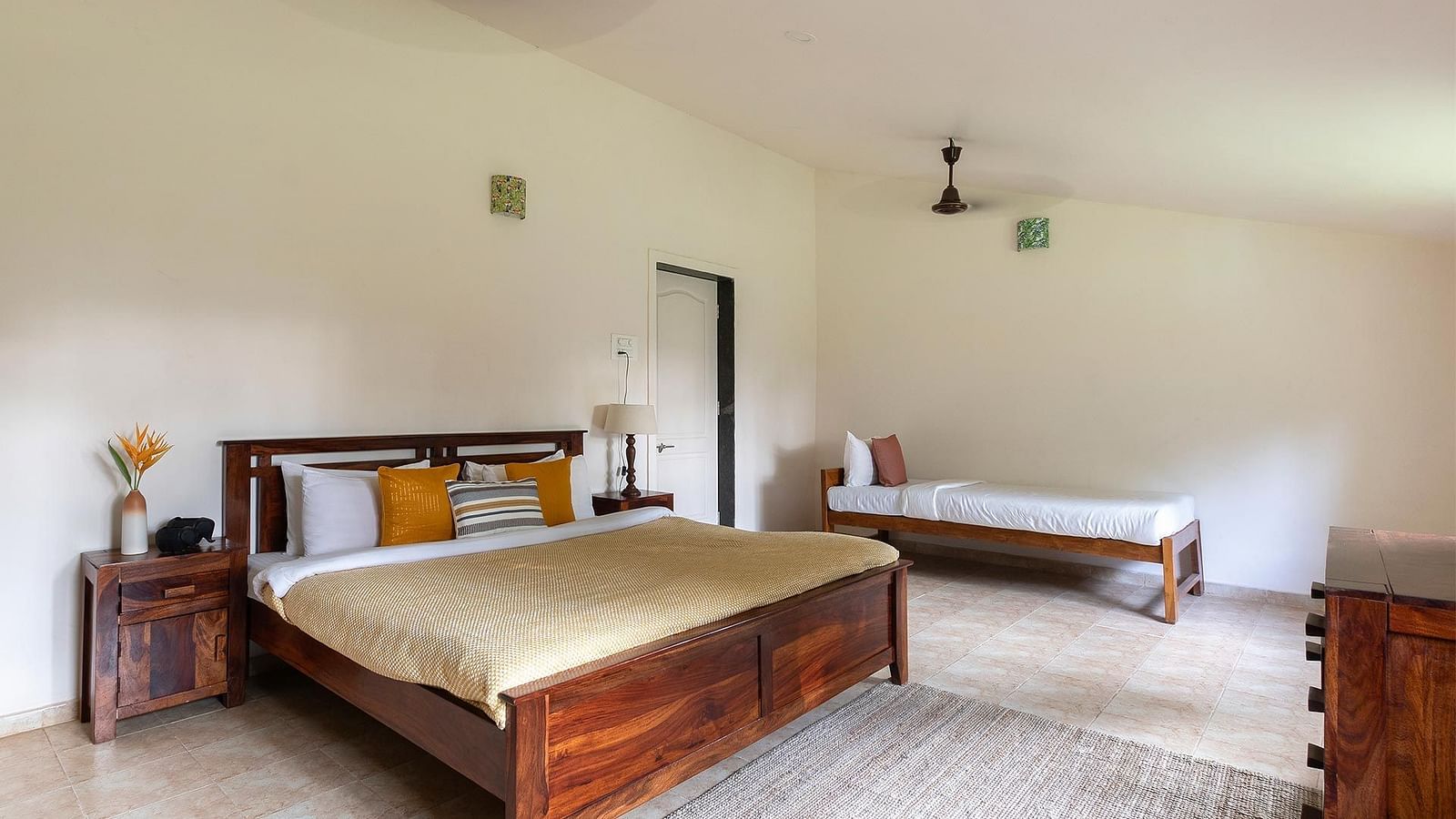 A side view of a double bed room with a single bed, beige carpet, ceiling fans, side tables and a door at The Conservancy Bungalow