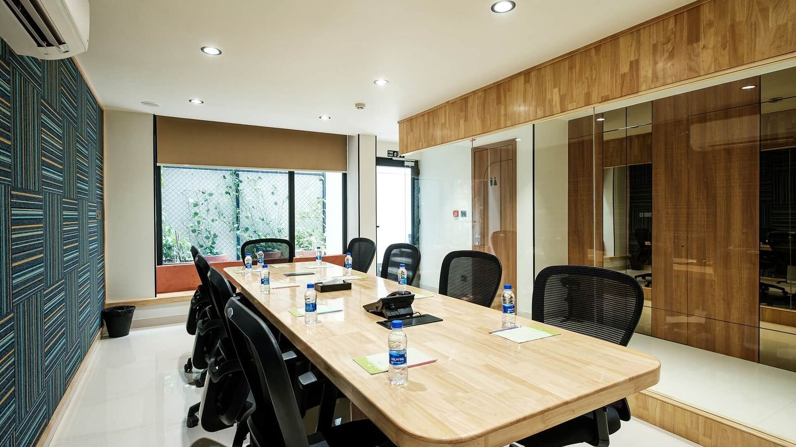 Diagonal view of the full boardroom at Indie Stays, Jaipur, with chairs arranged along the sides, a wall-mounted AC, and large windows at the end.