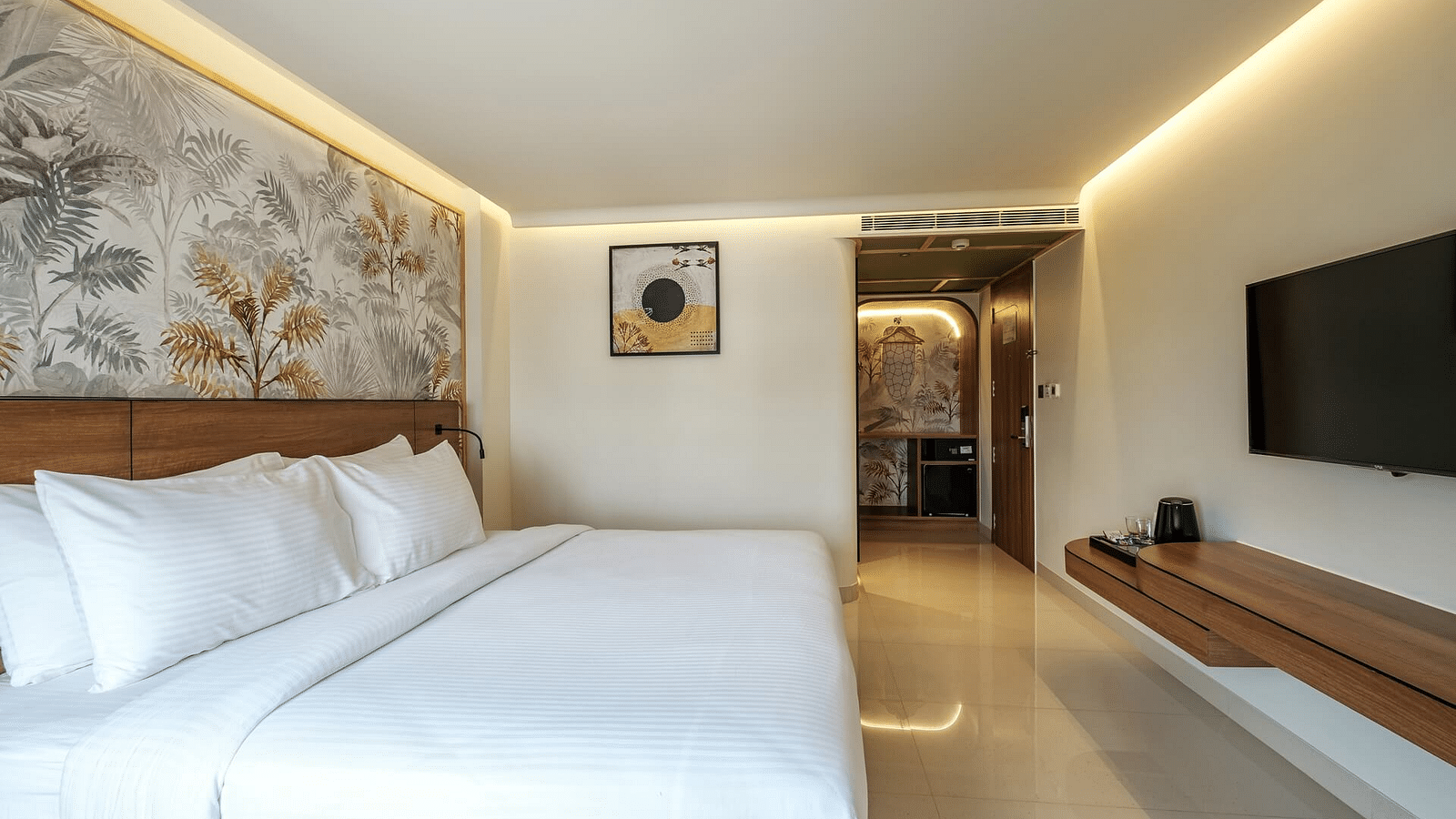 A side view of a spacious room at Indie Stays, Jaipur, featuring a large bed with a wooden headboard, a wall-mounted TV, and a pathway to the entrance.