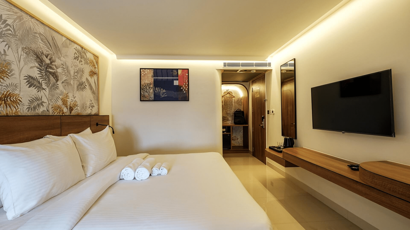 A side view of a spacious room at Indie Stays, Jaipur, featuring a large bed with a wooden headboard, a wall-mounted TV, and a pathway to the entrance.
