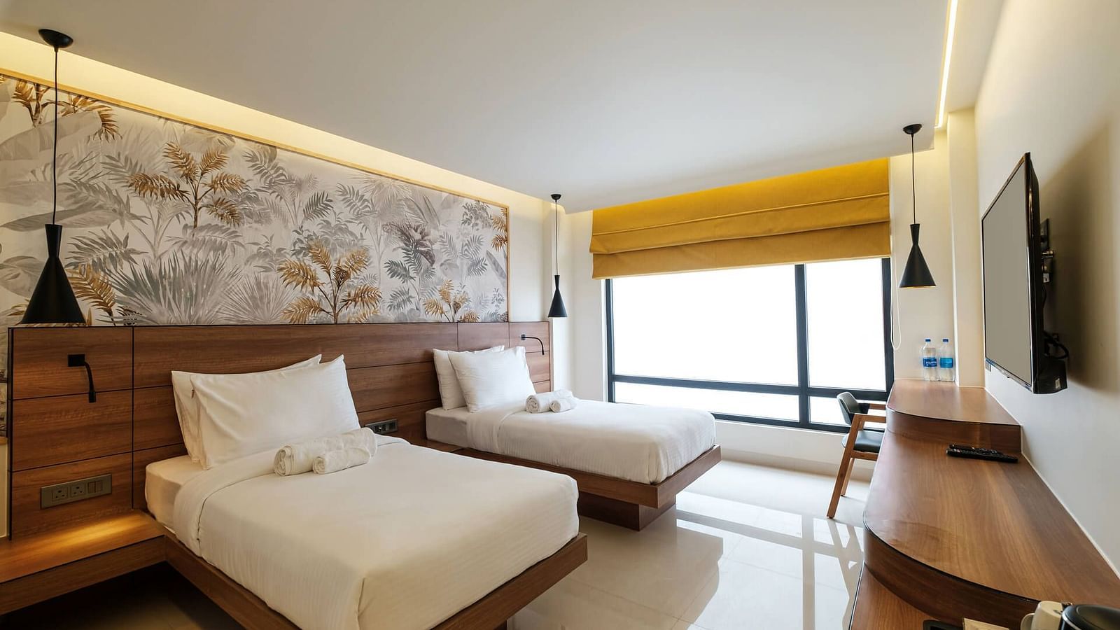 A spacious room at Indie Stays, Jaipur, with two beds and a joined wooden headboard, wall-mounted TV, wooden desk with a chair, and large alcove windows.