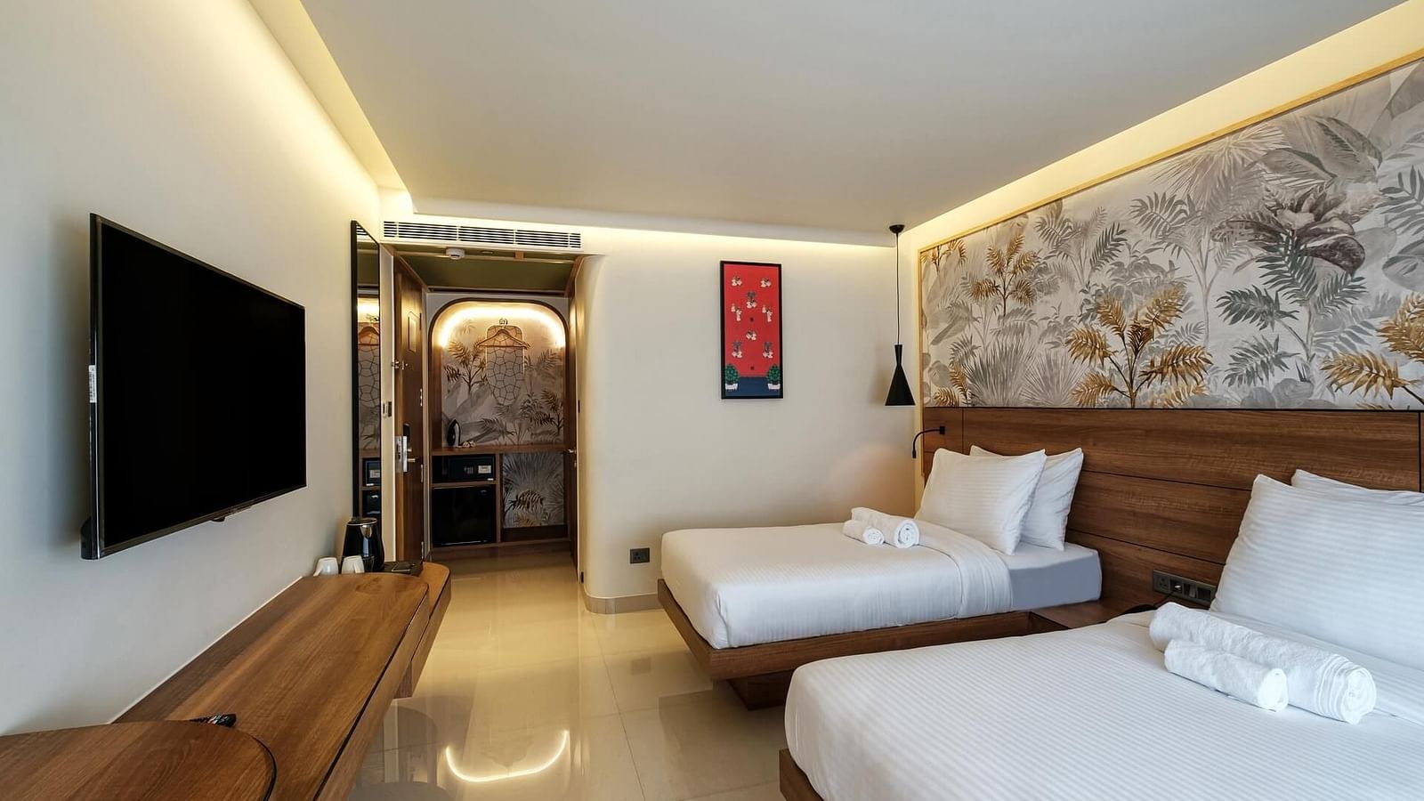 A side view of a spacious room at Indie Stays, Jaipur, with two single beds, wooden headboards, a wall-mounted TV with a wooden shelf below, and a pathway leading to the entrance.