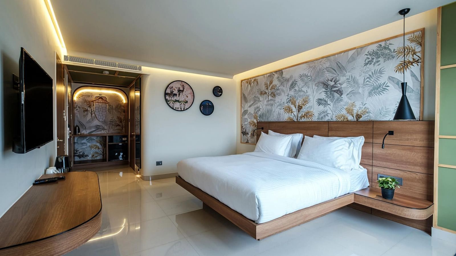 Spacious room at Indie Stays, Jaipur, featuring a large bed with a wooden headboard, a wall-mounted TV, and a pathway to the entrance.