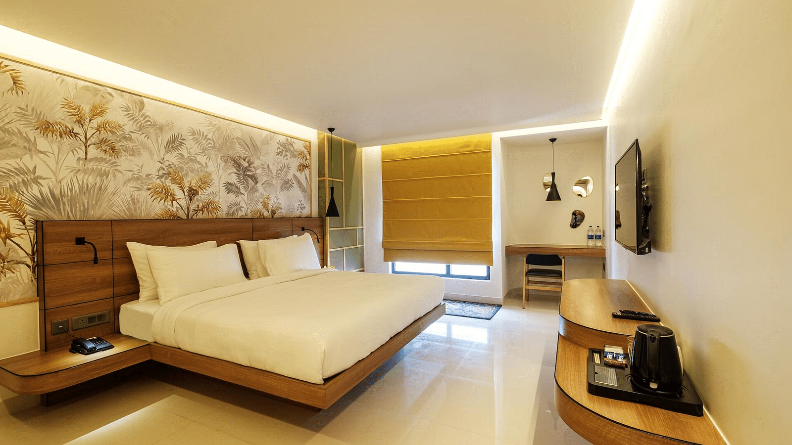 A spacious room at Indie Stays, Jaipur, featuring a large bed with a wooden headboard, a wall-mounted TV with a wooden shelf below, and curtained windows.
