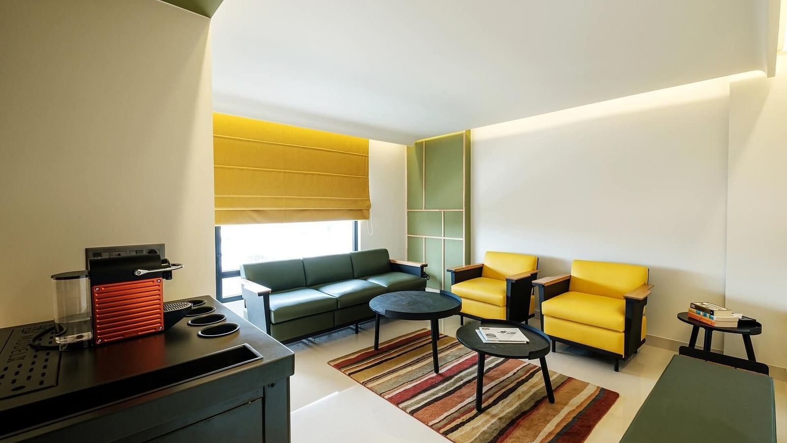Large cushioned sofas and two round coffee tables placed in the centre, above a floor mat, with curtains pulled halfway down and a small storage unit to the side at Indie Stays, Jaipur.