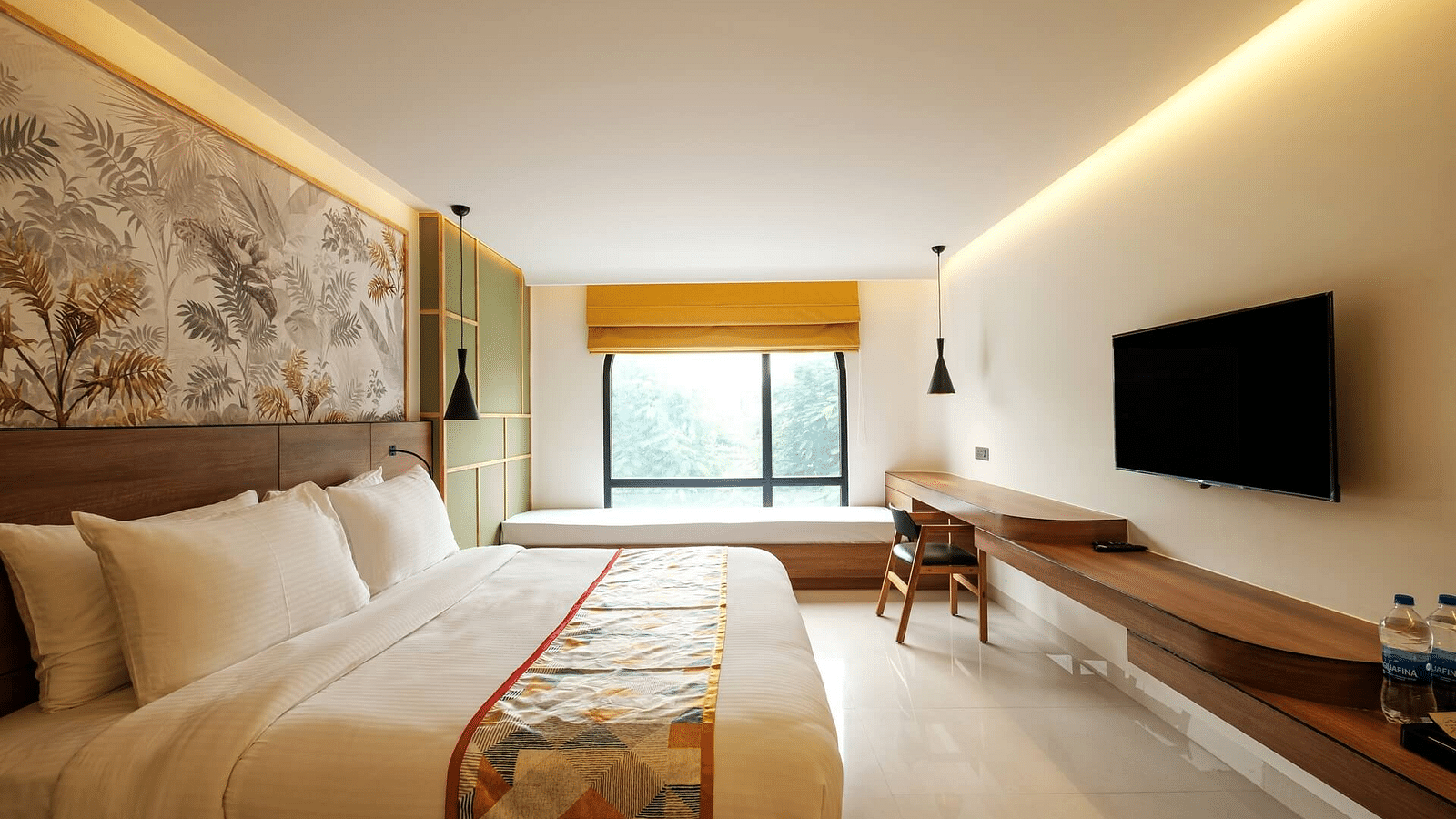 A side view of a spacious room at Indie Stays, Jaipur, featuring a large bed with a wooden headboard, a wall-mounted TV with a wooden shelf below, and curtained windows.