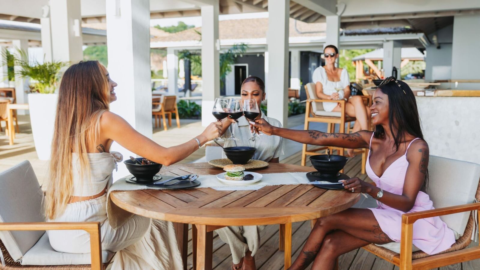  Three women toast with drinks at an outdoor restaurant, enjoying a meal under a shaded patio.