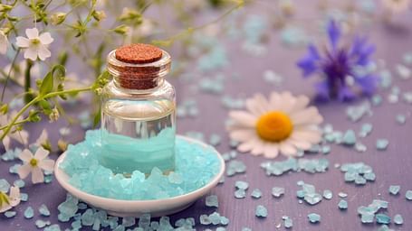 A small glass bottle with a cork top filled with essential oil, placed in a bowl of blue bath salts, surrounded by scattered salts, chamomile flowers, and delicate wildflowers, creating a calming spa-like setting