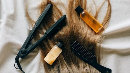 Long hair spread out on a cloth surface with a hair straightener, comb, and bottles of shampoo and conditioner placed on top