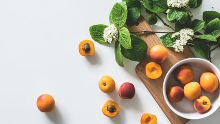 A bowl of peaches with some peaches outside, next to flowers with green leaves