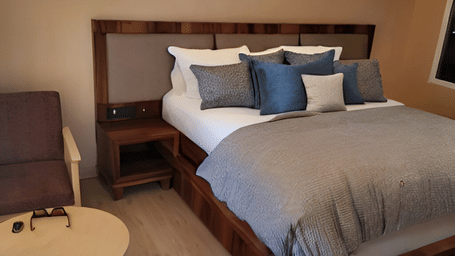 A close-up of a Deluxe Room at Hotel Dekiling by Eastlynn Collections, Leh with a wooden headboard and decorative pillows placed on a double bed with a bedside table and seating area right beside.