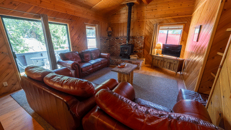 A living room with wood-paneled walls and vaulted ceiling, featuring brown leather sofas, a brick fireplace with wood stove, wooden coffee table, TV unit, and large windows with tree views - Shaver Lake Village Hotel