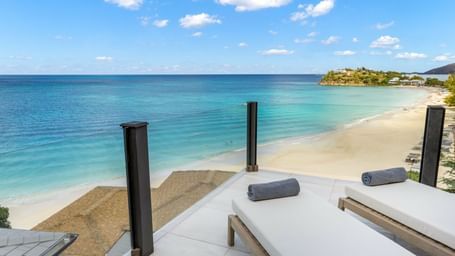A stunning view from a modern balcony with glass railings, overlooking a pristine white sand beach, turquoise ocean, and distant coastline under a blue sky. Two lounge chairs with rolled towels are on the tiled balcony.