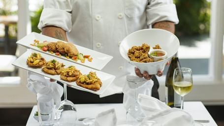 A person holding two plates and a bowl of seafood behind a table with neatly arranged cutlery at Tamarind Hills Resort and Villas, Antigua.
