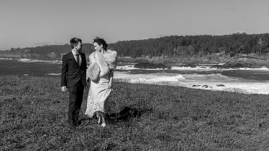 A black-and-white image of a couple walking near the ocean on a breezy day
