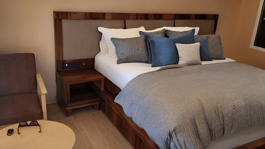 A close-up of a Deluxe Room at Hotel Dekiling by Eastlynn Collections, Leh with a wooden headboard and decorative pillows placed on a double bed with a bedside table and seating area right beside.