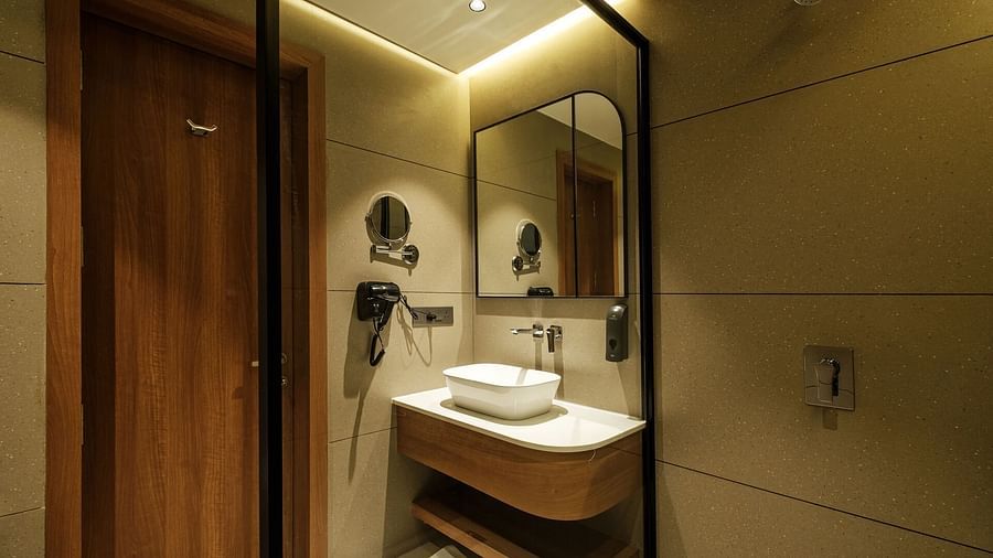 A spacious bathroom at Indie Stays, Jaipur, with a wooden door, a large wall-mounted mirror above the wash basin, and a hairdryer and handwash dispenser to the side.