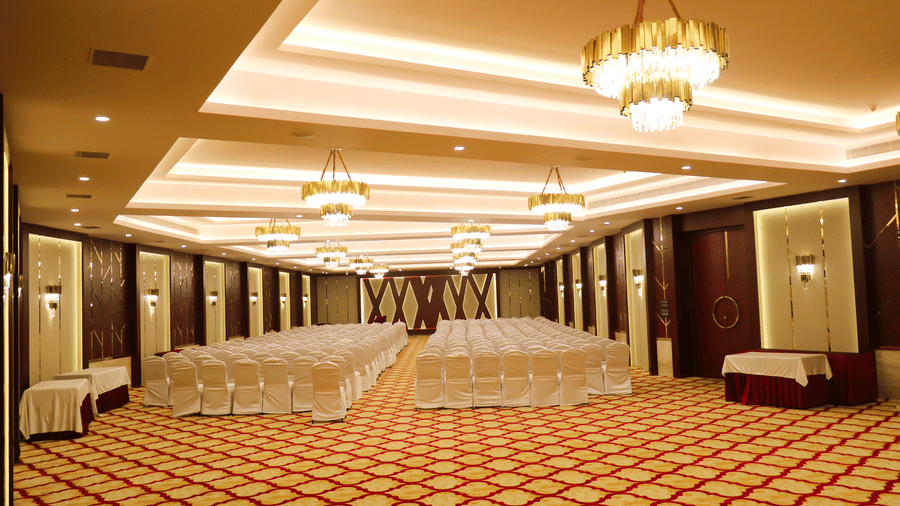 Spacious, elegant banquet hall at Jenneys Residency with patterned carpet, rows of chairs covered in cloth, decorative chandeliers, and warm lighting, set up for an event or conference