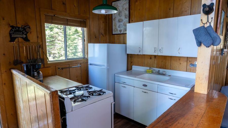 Small kitchen with stove, white cabinets, and window views in a forest cabin at Kit Carson Lodge.