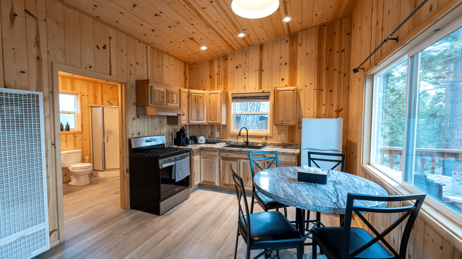 A kitchenette and a dining area at Kit Carson Lodge with a dining table, chairs, refrigerator, stove, and cabinets, surrounded by wooden walls and large windows offering forest views