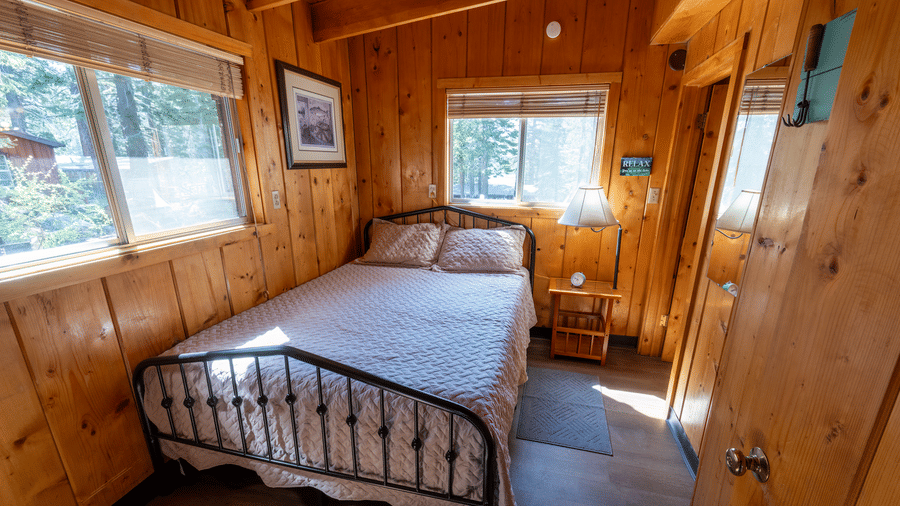 A bedroom at Kit Carson Lodge featuring a double bed, wooden walls, bedside tables, and windows offering views of the surrounding forest