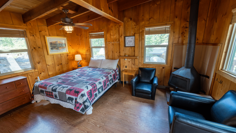 A bedroom at Kit Carson Lodge with a double bed,  2 armchairs, a side table, and large windows bringing in forest views