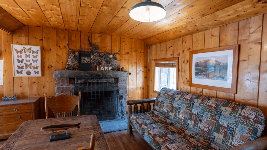 A living area at Kit Carson Lodge with a sofa, wooden interior, framed artwork, and a stone fireplace creating a rustic cabin atmosphere
