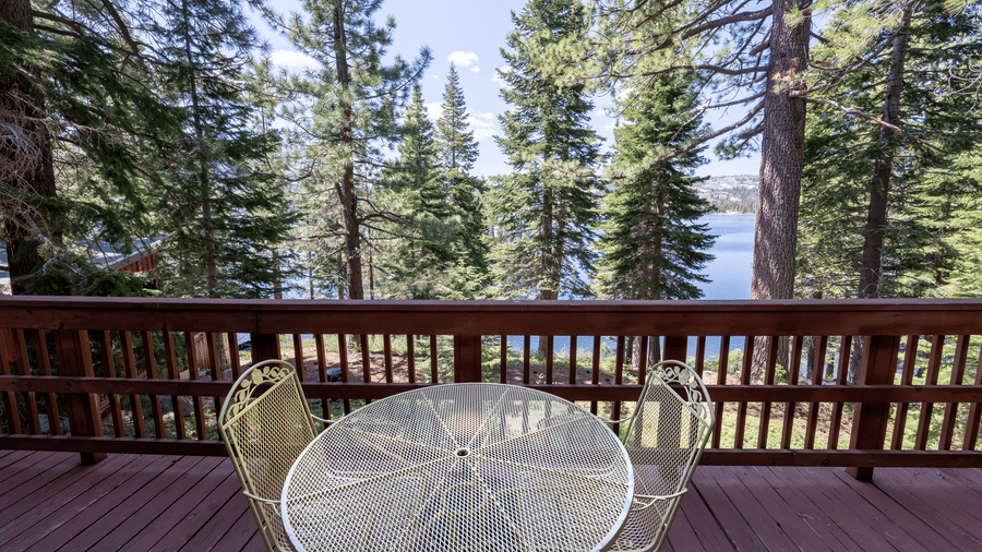 An outdoor deck at Kit Carson Lodge with a round table and chairs offering a view of the forest and lake through tall pine trees