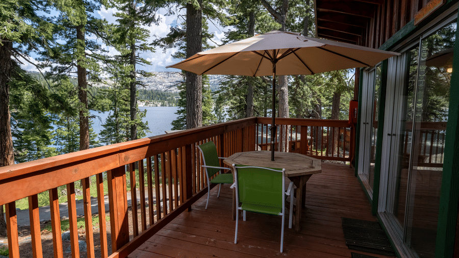 An outdoor deck at Kit Carson Lodge featuring a table with chairs and a large umbrella, overlooking tall pine trees and the nearby lake
