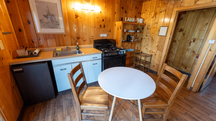A kitchenette at Kit Carson Lodge featuring a round dining table with chairs, compact refrigerator, stove, and cabinets against wood-paneled walls