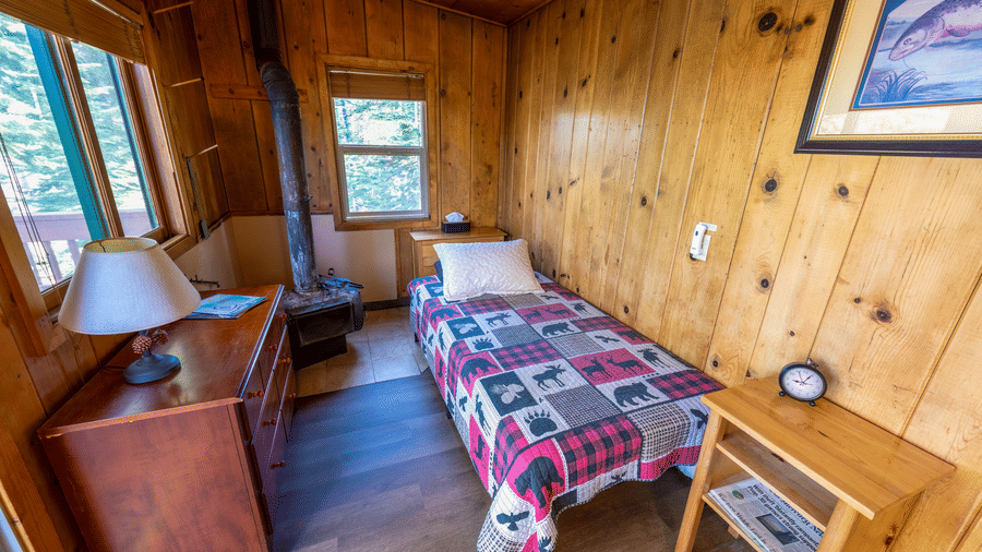 A single bedroom at Kit Carson Lodge with a neatly arranged bed, wooden walls, and a bedside table with a lamp beside the window