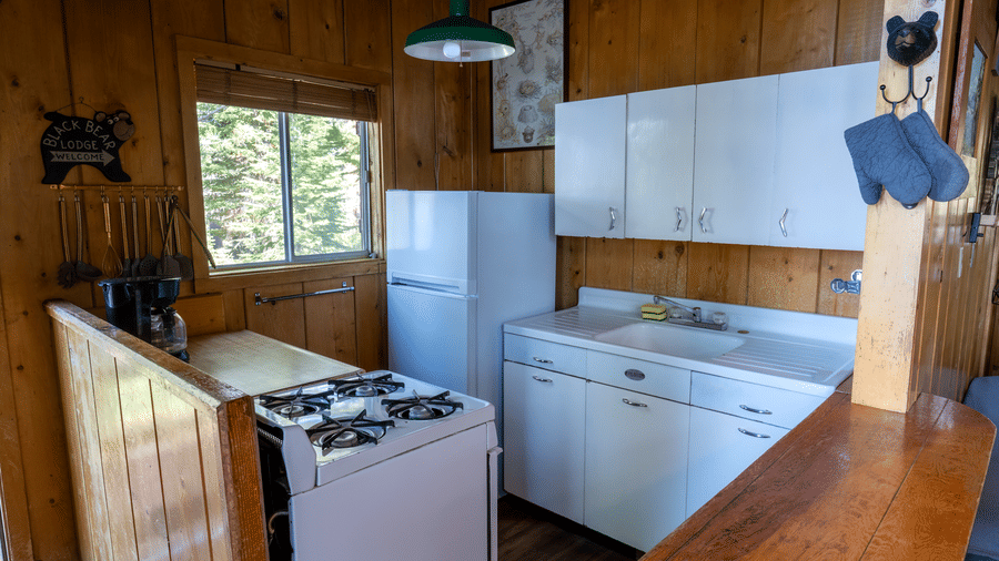 Small kitchen with stove, white cabinets, and window views in a forest cabin at Kit Carson Lodge