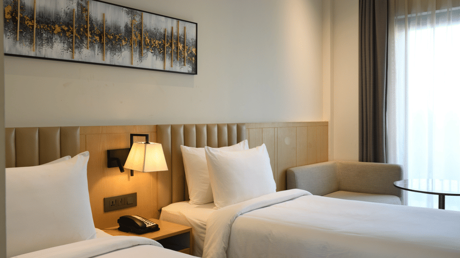 Neatly arranged twin-bed room with white bedding, a classic table lamp, wooden headboard, sheer curtained windows, and wall art above.