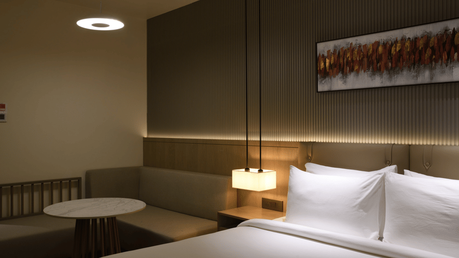 Cosy hotel room corner with a neatly made bed, pendant bedside lighting, warm wooden accents, and a vibrant abstract wall artwork.