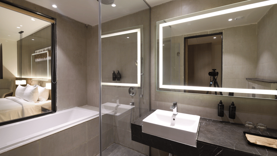 Modern bathroom at Ramada by Wyndham Sonipat Murthal, featuring dual backlit vanity mirrors, a sleek countertop basin, and a glass-enclosed shower with grey tile walls.
