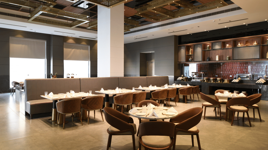 Spacious all-day dining restaurant with round tables, cushioned chairs, and modern gold shelving units against large windows.