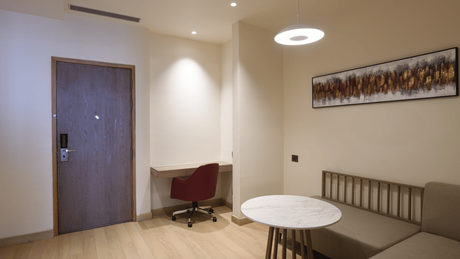 Hotel room entryway at Ramada by Wyndham Sonipat Murthal, featuring a wooden door, a round white table, a chair, and soft recessed ceiling lighting.