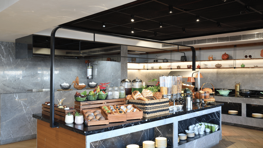 Modern breakfast buffet island with marble counters, featuring tiered displays of fresh pastries, breads, cereals, and fruits | Ramada Encore by Wyndham Gurugram Dwarka Expressway