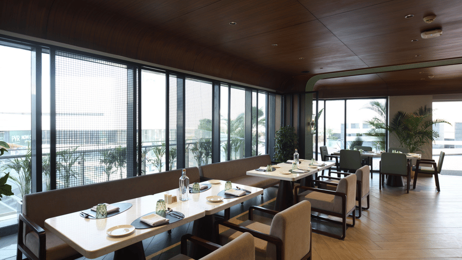 Dining space with white tables, wood-panelled ceilings, and mesh-covered windows overlooking the buildings outside | Ramada Encore by Wyndham Gurugram Dwarka Expressway