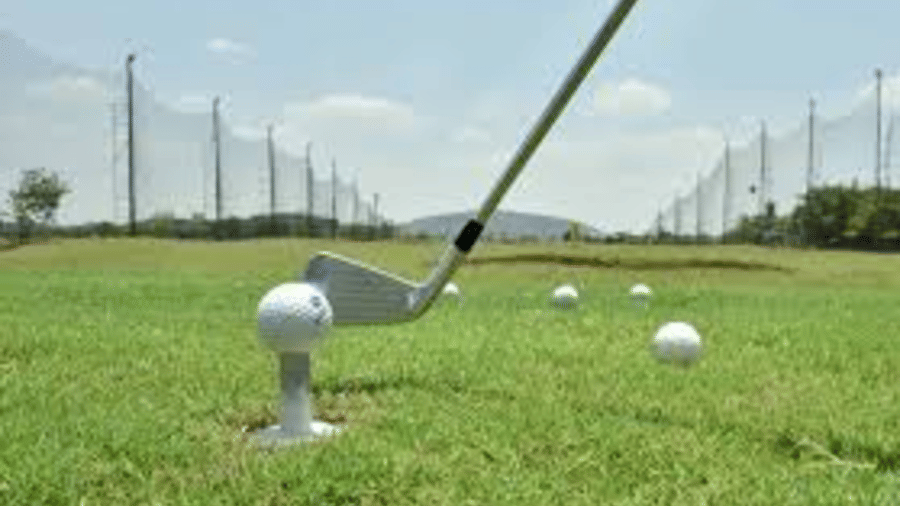 A close-up shot of a golf iron about to hit a ball on a tee at a driving range, with protective netting and green fields in the background at Apartel by Aarin, Oragadam.