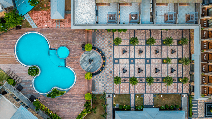 An aerial view of the swimming pool of Pushkara Resort & Spa, Ajmer, one of the Best Resorts in Ajmer, with tables set beside it on the stone flooring.
