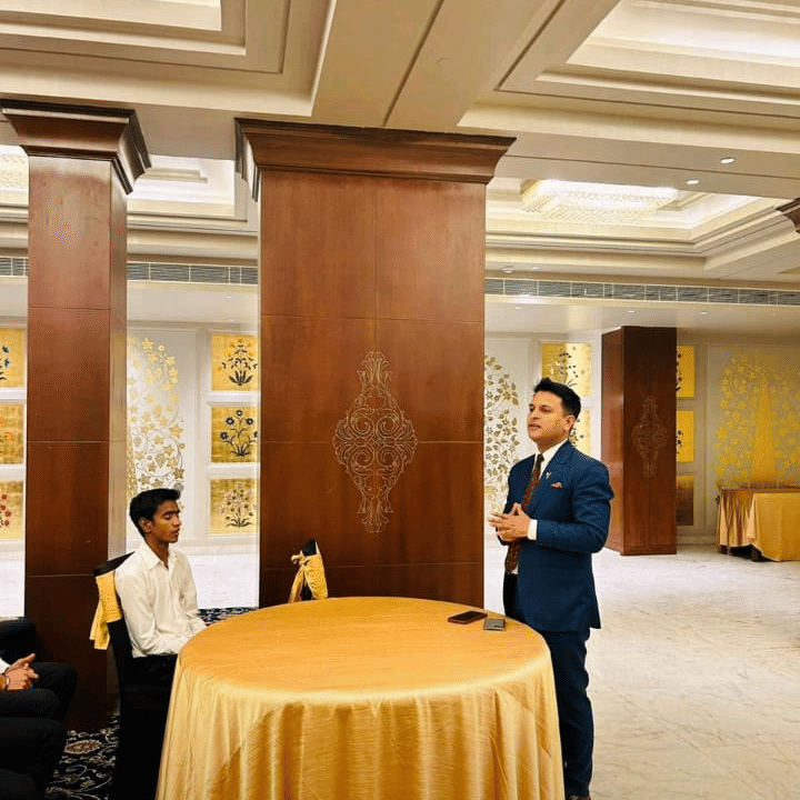 A speaker addresses an audience inside a hall at Clarks Group of Hotels. A table stands next to the presenter.