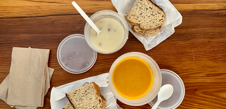 Two containers of soup, one light and one dark, sit on a wooden table next to two pieces of bread and napkins.