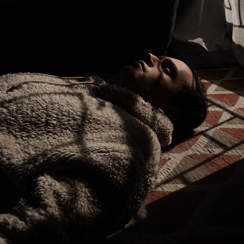 A man lies on a patterned rug, partially covered by a blanket, with sunlight casting shadows on him.