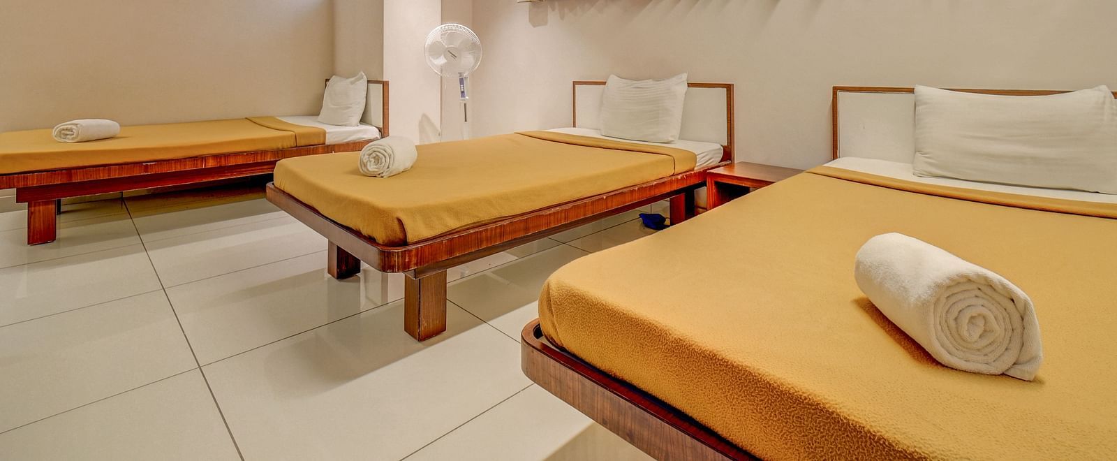A spacious room with three neatly made beds, each with rolled towels placed over the blanket, and a rectangular window panel covered with curtains at Harigovinda's JNStay