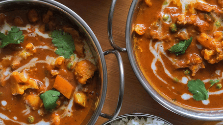 A top-down view of two metal bowls of curry garnished with herbs served alongside two bowls of white rice on a wooden surface.